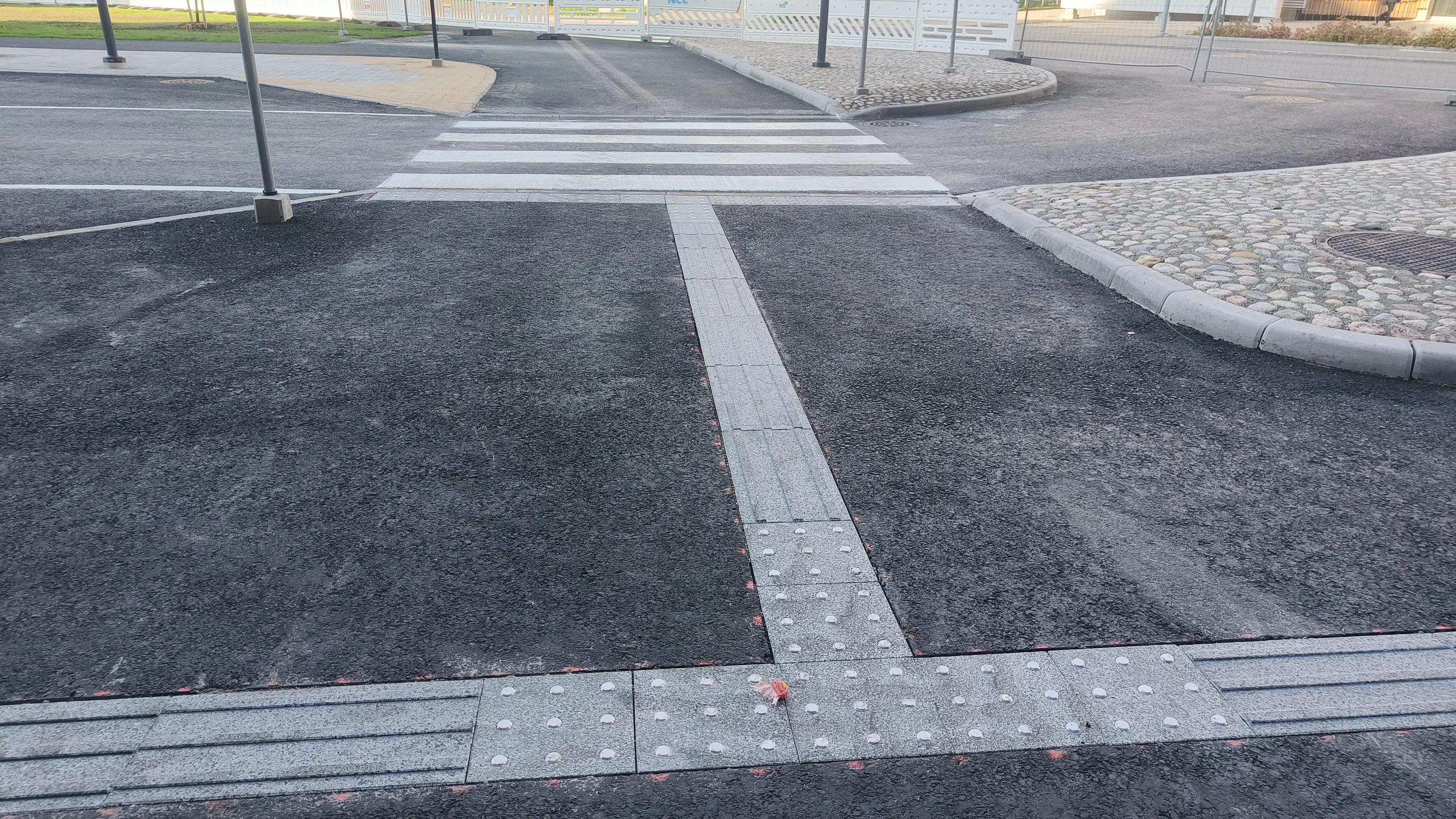Kuva esittää vastapäällystettyä tummanharmaata asfalttitietä. Etualalla on L-muotoinen vaaleanharmaa tuntoesteetön laatoitus, jossa on sekä kohoviivoja että kohopisteitä. Laatoituksen kulmassa on pieni, värikäs roskanpala. Keskellä kuvaa on valkoinen suojatie. Oikealla puolella kaareva reunakivi erottaa asfaltin mukulakivialueesta, jossa on pyöreä kaivonkansi. Taustalla näkyy lisää asfalttia, vaaleaa päällystettä, valkoisia rakennusaitoja, vihreää ruohoa ja tummia pylväitä. Pinnat näyttävät kosteilta.