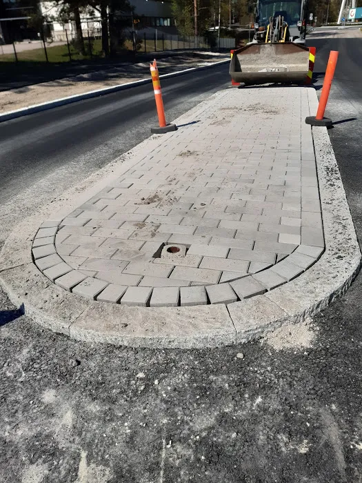 Vaaleanharmaa kiveyssaareke on keskellä mustaa asfalttitietä aurinkoisena päivänä. Saarekkeen reunat ovat tummanharmaata reunakiveä, ja sen pinnassa on pieni pyöreä aukko. Kaksi oranssia liikennekartiota seisoo saarekkeen päällä. Taustalla näkyy keltainen työkone, puita ja rakennuksia.