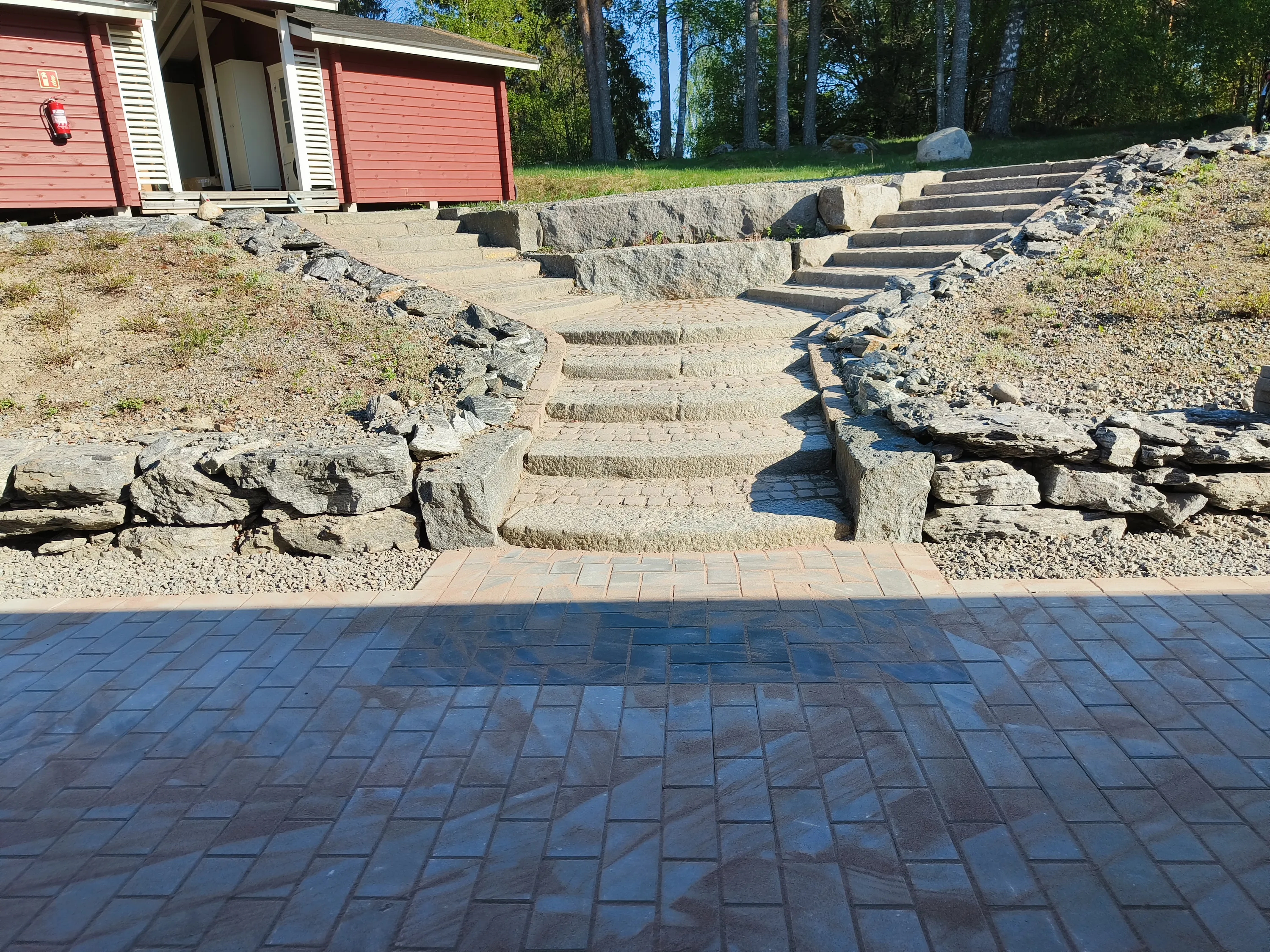 Aurinkoisena päivänä otetussa kuvassa on etualalla tummanharmaista laatoista tehty polku, jossa näkyy varjoja. Keskellä nousevat vaaleanharmaat kiviset portaat, jotka haarautuvat kahteen suuntaan. Portaiden ja polkujen reunoilla on kivimuureja. Vasemmalla taustalla on punainen puurakennus valkoisine ikkunaluukkuineen ja seinällä olevine palosammuttimineen. Oikealla taustalla on vihreä nurmikko ja puita sinistä taivasta vasten.