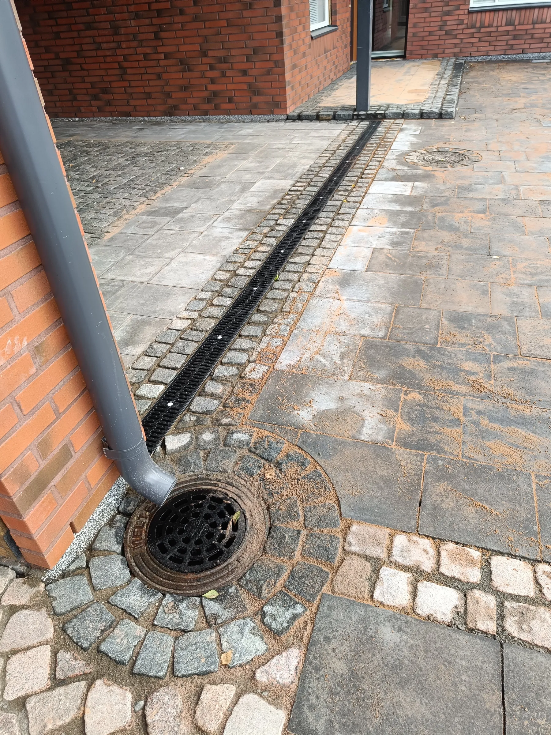 Kuvassa on tiilirakennuksen vieressä oleva kivetty piha. Harmaa syöksytorvi johtaa mustaan pyöreään kaivonkanteen. Pihalla kulkee pitkä musta linjakuivatuskouru, jota reunustavat harmaat nupukivet. Muu laatoitus koostuu harmaista suorakaiteen muotoisista pihakivistä. Kivetysten päällä on siroteltuna vaaleaa hiekkaa.