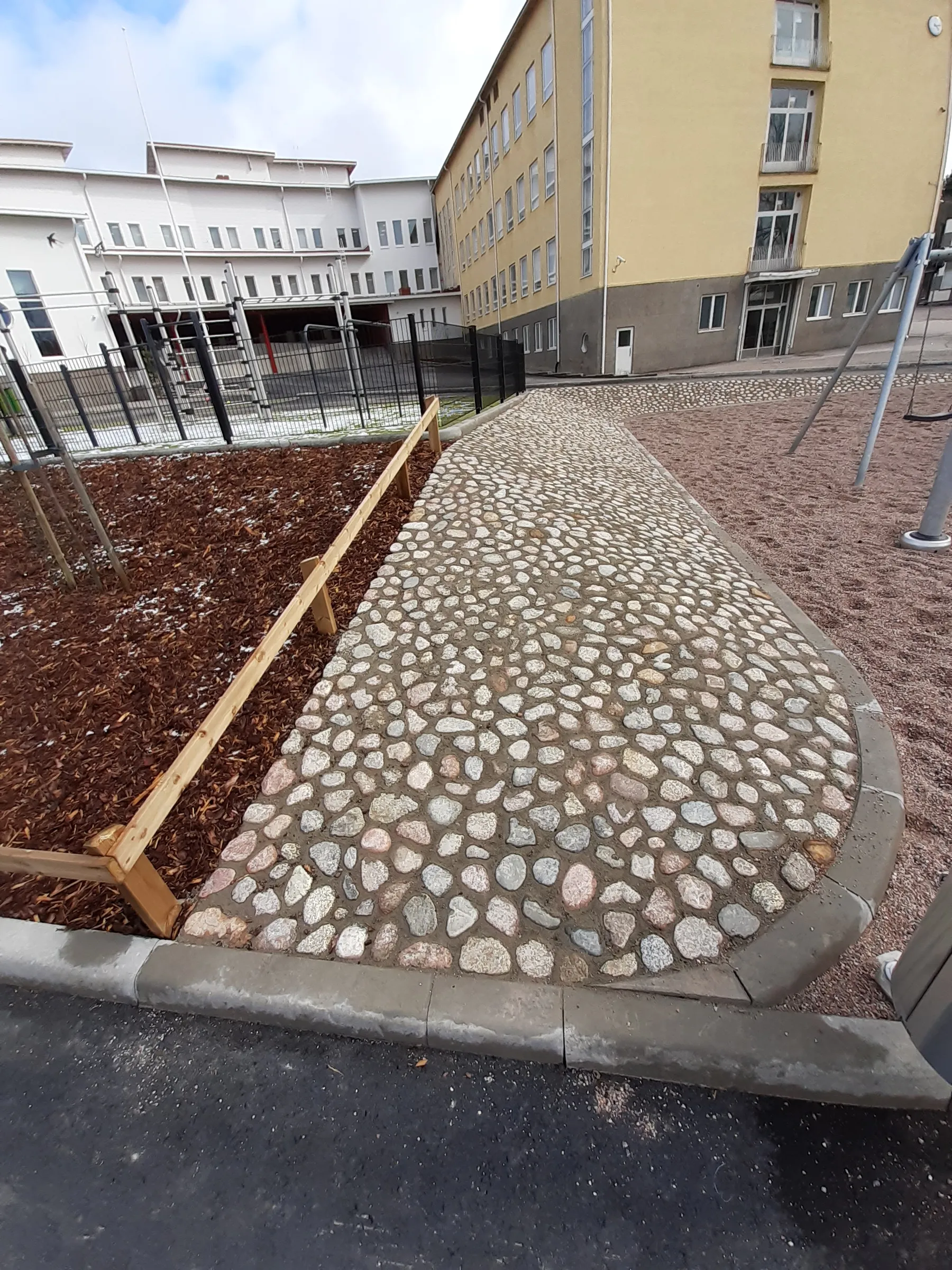 Etualalla on tumma asfalttitie, jonka vieressä on ruskeaa puuhaketta ja pieniä lumiläiskiä. Puuhaketta rajaa vaalea puureunus. Keskellä on kaareva, vaaleista kivistä ja tummasta laastista ladottu polku. Polun oikealla puolella on punertavanruskea leikkipaikan alusta, jossa on keinutelineen tolppa. Taustalla näkyy valkoinen rakennus mustan aidan takana ja keltainen rakennus harmaalla alaosalla. Pilvinen taivas on vaaleansininen.