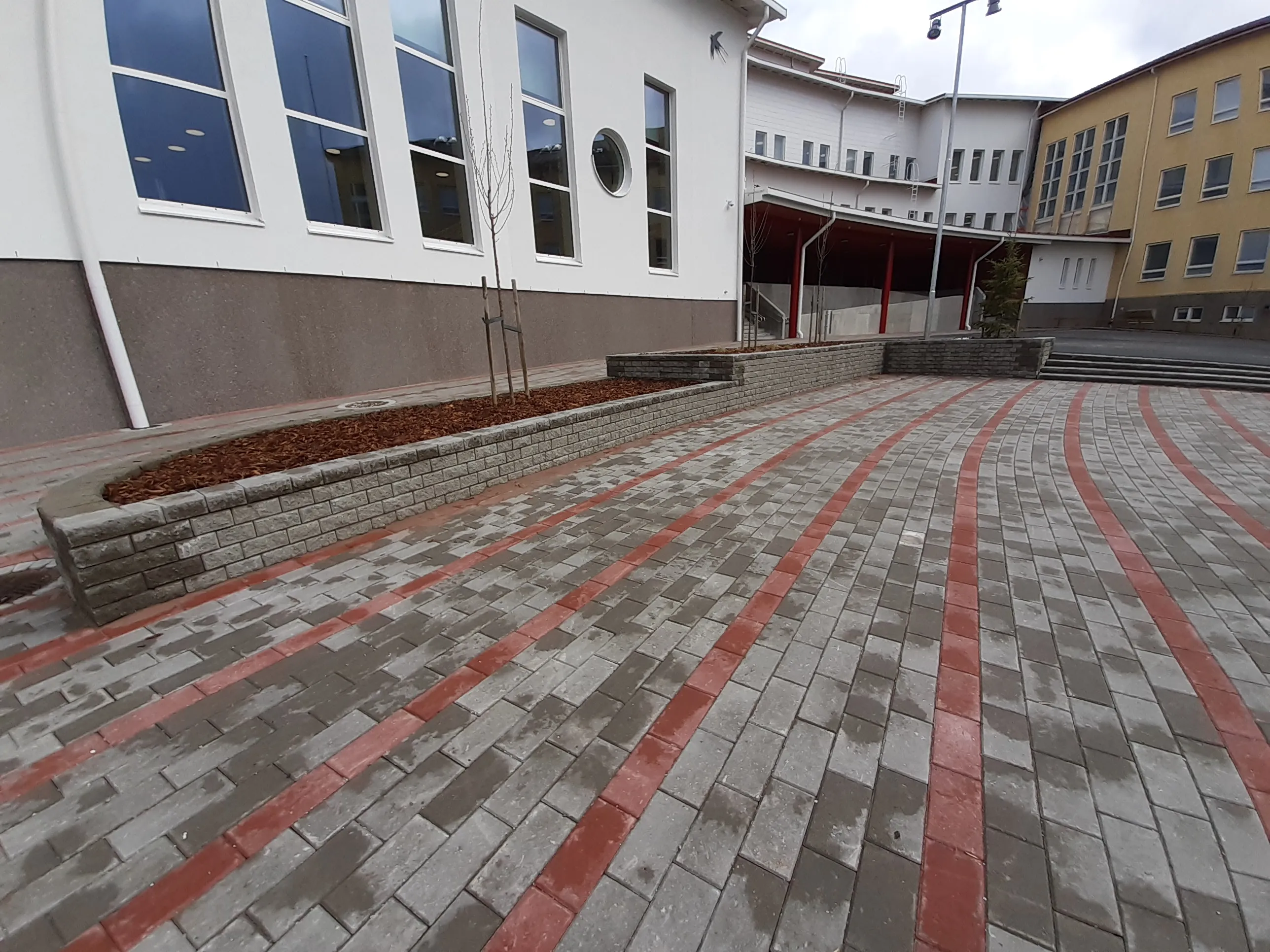 Kuvassa on kivetty piha-alue useiden rakennusten edessä pilvisenä päivänä. Pihan harmaassa ja tummanharmaassa kivetyksessä on kaarevia punaisia raitoja. Vasemmalla on valkoinen rakennus, jossa on suuria ja yksi pyöreä ikkuna, sekä harmaa alaosa. Sen edessä on harmaista tiilistä tehty istutuslaatikko, jossa on ruskeaa kuorikatetta ja nuoria puita. Taustalla näkyy valkoinen rakennus punaisella katoksella ja keltainen rakennus portaineen. Kivetys on märkä.