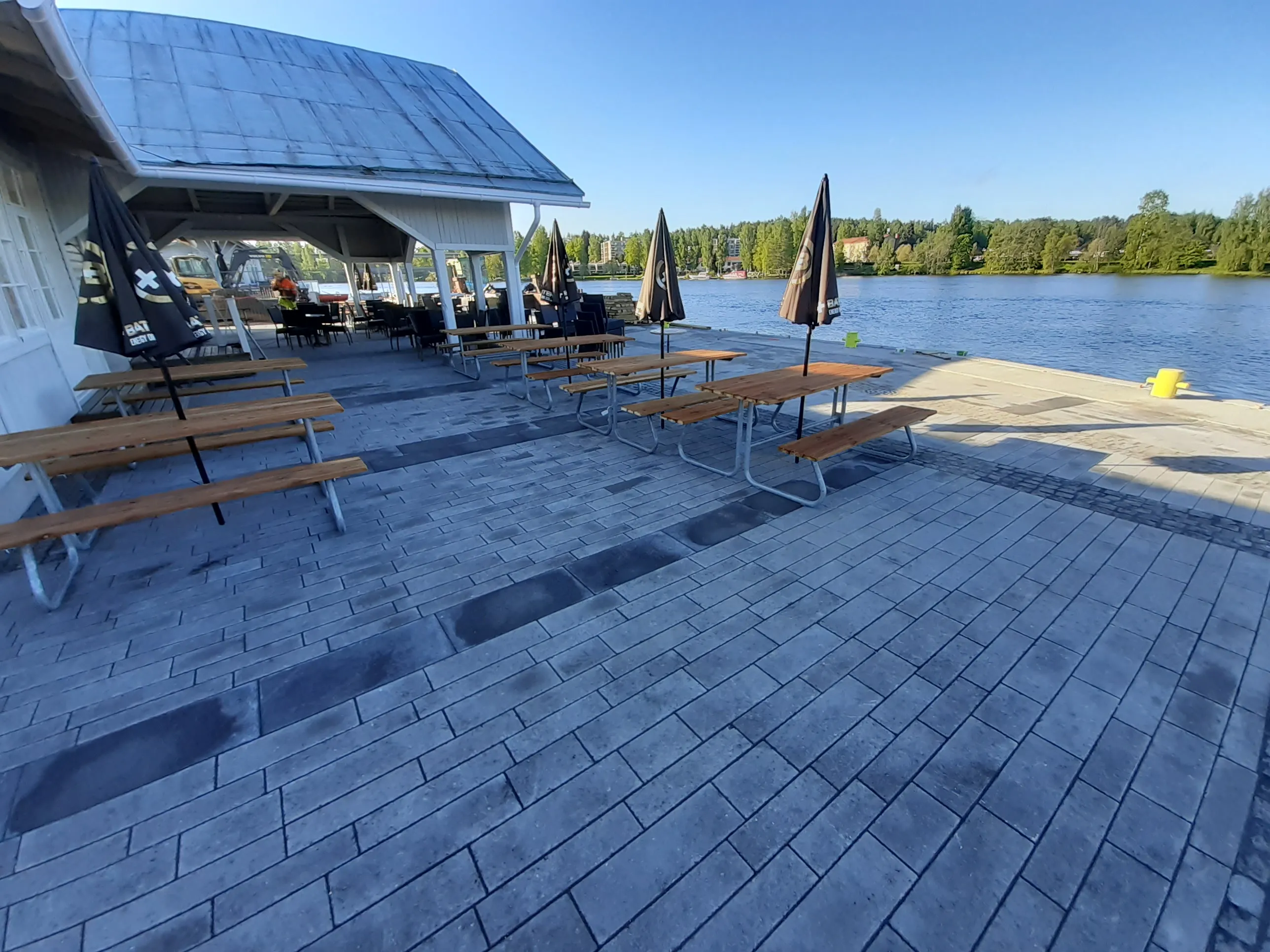 Aurinkoinen ulkokuva joen tai järven rannalla sijaitsevasta kahvilasta. Harmaalla laatoituksella on useita ruskeita puupiknikpöytiä ja mustia suljettuja aurinkovarjoja. Vasemmalla näkyy valkoisen rakennuksen osa harmaalla katolla. Laiturin reunalla on keltainen pollari. Taustalla on sininen vesi, jonka vastarannalla on vihreä puurivi ja rakennuksia. Kirkas sininen taivas ja pitkät varjot laatoituksella.