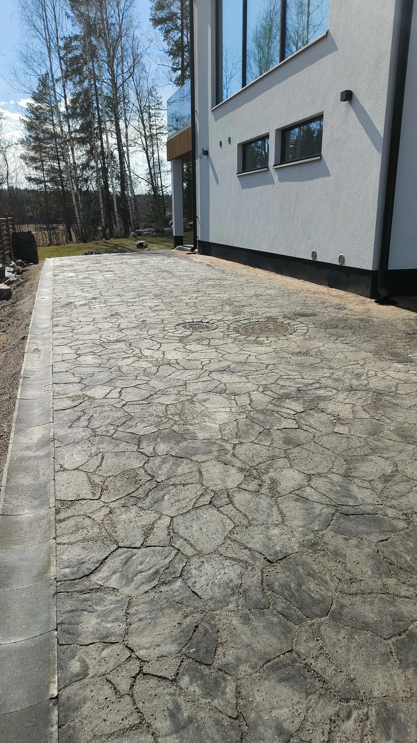 Aurinkoisena päivänä otettu kuva modernista valkoisesta talosta ja sen vieressä olevasta kivetystä piha-alueesta. Piha on päällystetty epäsäännöllisen muotoisilla harmailla pihakivillä, joiden saumoissa on hiekkaa. Pihan reunalla on sadevesikouru ja kaksi pyöreää kaivonkantta. Talon valkoisessa seinässä on suuria heijastavia ikkunoita ja kaksi pienempää ikkunaa. Taustalla näkyy metsää ja avoin maisema sinisen taivaan alla.