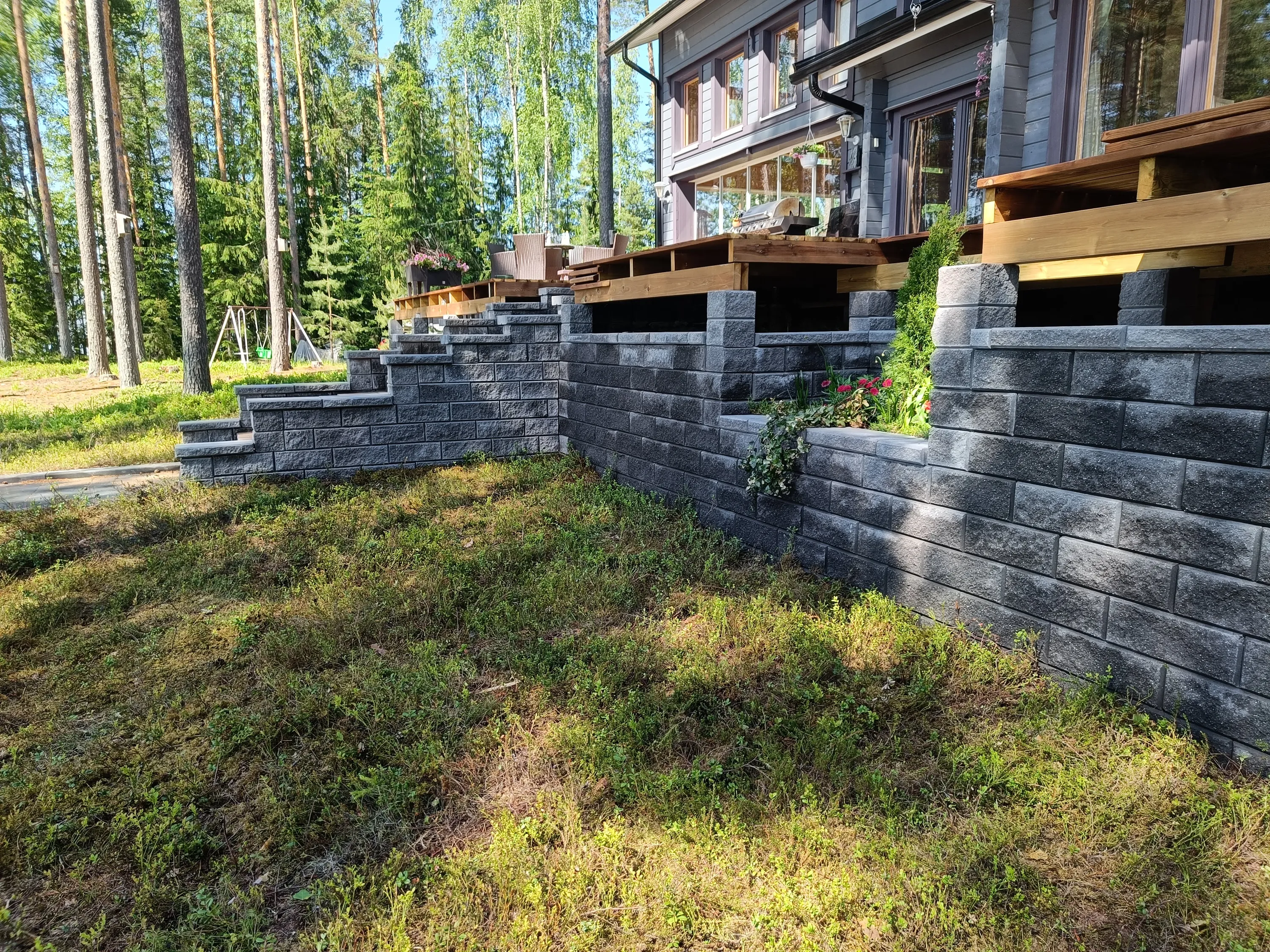 Aurinkoinen kuva harmaasta talosta, jossa on puinen terassi ja harmaa kivimuuri portaineen. Terassilla on ulkokalusteita ja grilli. Taustalla on tiheä metsä, jossa on mäntyjä ja koivuja. Etualalla maata peittää vihreä ja ruskehtava varpukasvillisuus. Muurissa on istutuksia ja puiden rungoissa linnunpönttöjä.